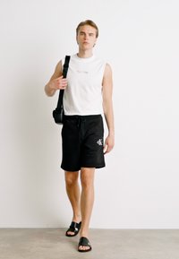 Camisa sin mangas blanca con texto multicolor, pantalones cortos negros con logo, sandalias negras, llevando una bolsa negra pequeña, de pie sobre un fondo neutro.