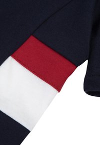 T-shirt en coton bleu marine avec des accents de rayures rouges et blanches sur les manches. Présente une texture lisse et des détails de couture soignés.