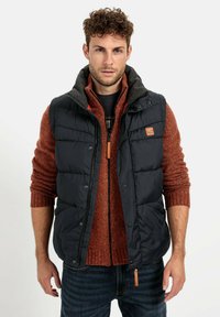 Marineblaue Daunenweste mit grauem Kragen über einem orangefarbenen Strickpullover. Verfügt über Reißverschluss- und Druckknopfverschlüsse sowie eine Lederakzent auf der Weste.