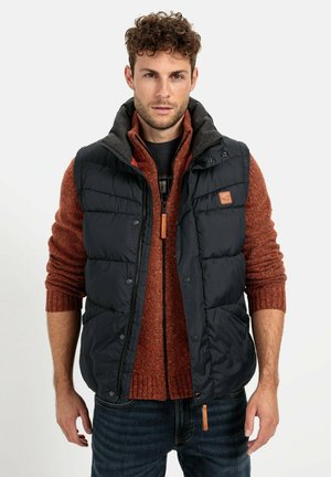 Marineblaue Daunenweste mit grauem Kragen über einem orangefarbenen Strickpullover. Verfügt über Reißverschluss- und Druckknopfverschlüsse sowie eine Lederakzent auf der Weste.