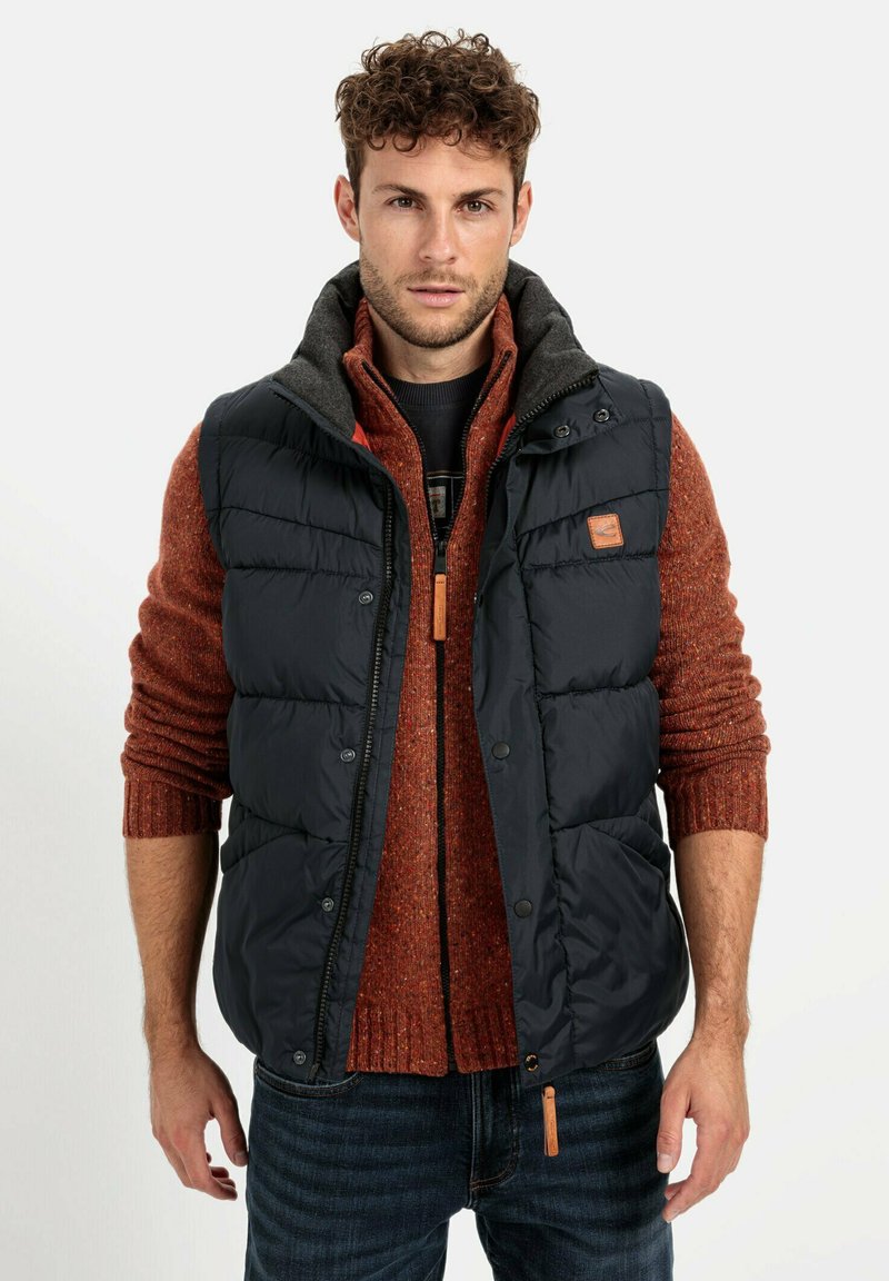 Marineblaue Daunenweste mit grauem Kragen über einem orangefarbenen Strickpullover. Verfügt über Reißverschluss- und Druckknopfverschlüsse sowie eine Lederakzent auf der Weste.