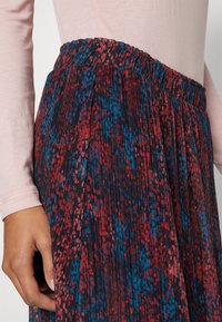 Jupe plissée avec une taille élastique, présentant une base sombre avec des motifs parsemés de rouge et de bleu. Texture lisse et silhouette fluide.