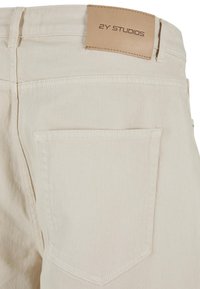Beige denim trousers featuring a back pocket and a tan leather label on the waistband with "2Y STUDIOS" embossed. Smooth texture.