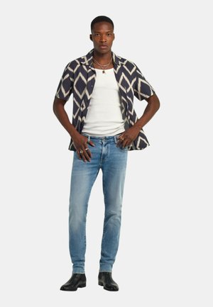 Giovane uomo in piedi con le mani sui fianchi, indossa una camicia a motivo diamante bianco e nero, una canotta bianca, jeans azzurri e stivali neri.