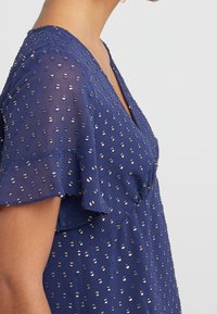 Blusa blu navy con maniche corte a volant, realizzata in un tessuto testurizzato decorato con motivi a pois metallici dorati e scollatura a V.