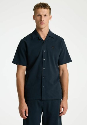 Fiatal férfi rövid, göndör hajjal, fekete rövid ujjú gombos ingben és hozzáillő fekete nadrágban, egyszerű fehér háttér előtt állva.