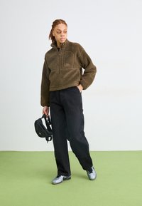 Brun fleece pullover med kvart glidelås, svarte cargo-bukser og sølvfargede joggesko. Personen holder en svart bag og står på en grønn matte.