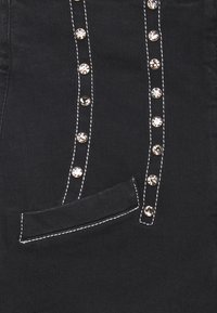 La tela de mezclilla negra presenta una línea vertical de clavos metálicos, un contraste de costuras blancas y un pequeño bolsillo de parche.