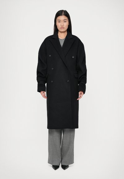 Woman stands wearing an oversized black double-breasted coat, gray pants, and black pointed shoes against a plain white background.