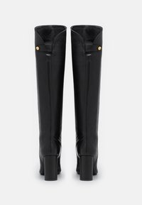 Bottes noires en cuir, montant jusqu'au genou, avec un talon large, présentant un design épuré et un bouton-pression doré en haut. Texture lisse.