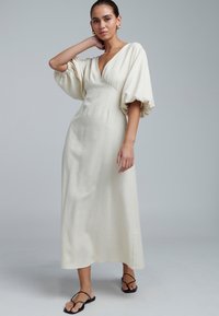 Ecrufarbenes Maxikleid mit tiefem V-Ausschnitt, luftigen Ärmeln und taillierter Passform, aus strukturiertem Stoff. Getragen mit schwarzen Flip-Flops.