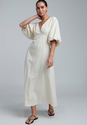 Cream-colored maxi dress with a deep V-neck, billowy sleeves, and a fitted bodice, made from textured fabric. Worn with black flip-flops.