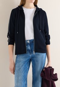 Personne portant un sweat à capuche bleu marine zippé sur un t-shirt blanc et un jean bleu, tenant un vêtement texturé plié de couleur violet foncé dans la main droite.