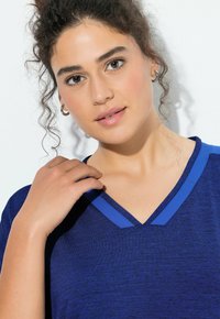 Młoda kobieta o kręconych włosach, nosząca srebrne kolczyki koła i niebieską koszulkę z dekoltem w serek, delikatnie dotykająca ręką linii dekoltu.