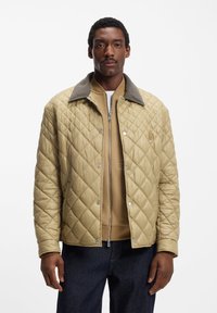 Κουβερτοειδές χακί μπουφάν με γκρι γιακά, κουμπιά με πατιλέτα και υφασμάτινη υφή, φορεμένο πάνω από μπεζ φούτερ και συνδυασμένο με σκούρο τζιν.