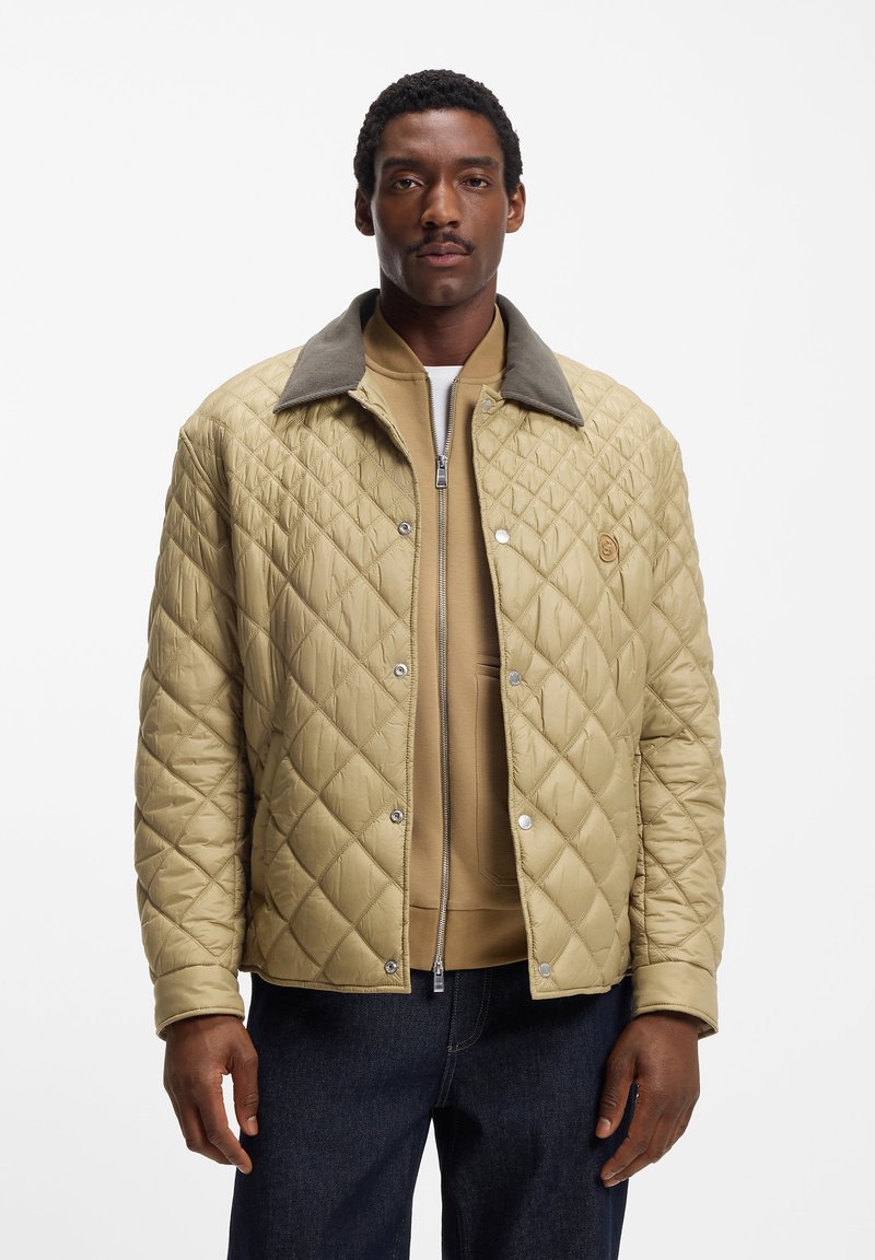 Κουβερτοειδές χακί μπουφάν με γκρι γιακά, κουμπιά με πατιλέτα και υφασμάτινη υφή, φορεμένο πάνω από μπεζ φούτερ και συνδυασμένο με σκούρο τζιν.