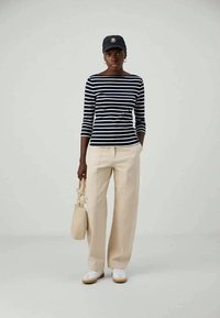 Marineblauw en witte gestreepte shirt met driekwart mouwen, beige wijde broek, witte sneakers en een zwarte pet, met een beige tas in de hand.