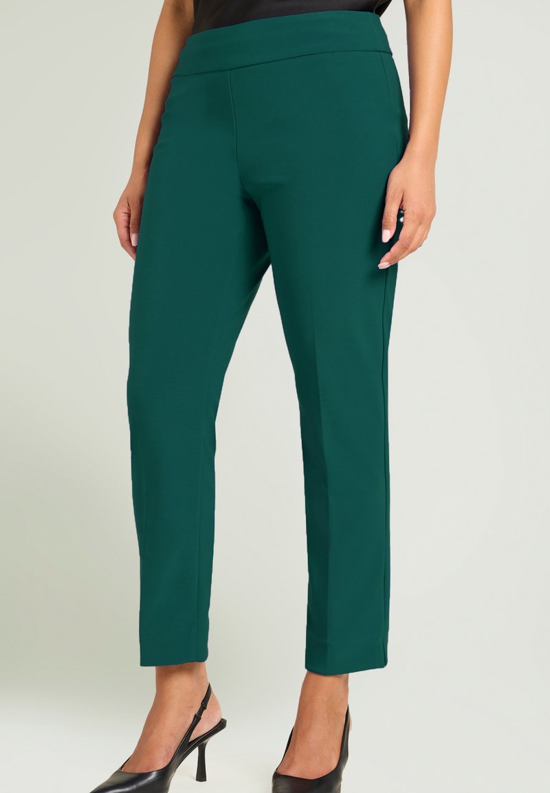 Groene, op maat gemaakte broek met een strak design, voorzien van een brede tailleband en rechte pijpen, gecombineerd met zwarte schoenen met hak.