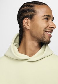 Svetlozelena kapuca z veliko mehko kapuco. Tkanina izgleda gladka in elastična, z oblikovanim ovratnikom. Vidne so pletene lase.