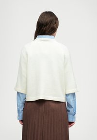 White cropped sweater with wide sleeves, textured fabric, paired with a light blue striped shirt and brown pleated skirt.
