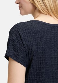 Maglietta blu navy con un tessuto testurizzato e sgualcito. Presenta maniche corte e un collo rotondo, mettendo in risalto elementi di design semplici.