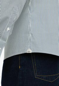 Blauses und weiße senkrechte gestreifte Hemd mit einem geschwungenen Saum. Das Etikett an der Seite zeigt "LIU-JO." Getragen mit dunklen Jeans.