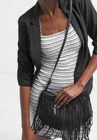 Vrouw in een zwarte blazer over een gestreepte jurk, met één hand een zwarte leren crossbody tas met franjes vasthoudend.