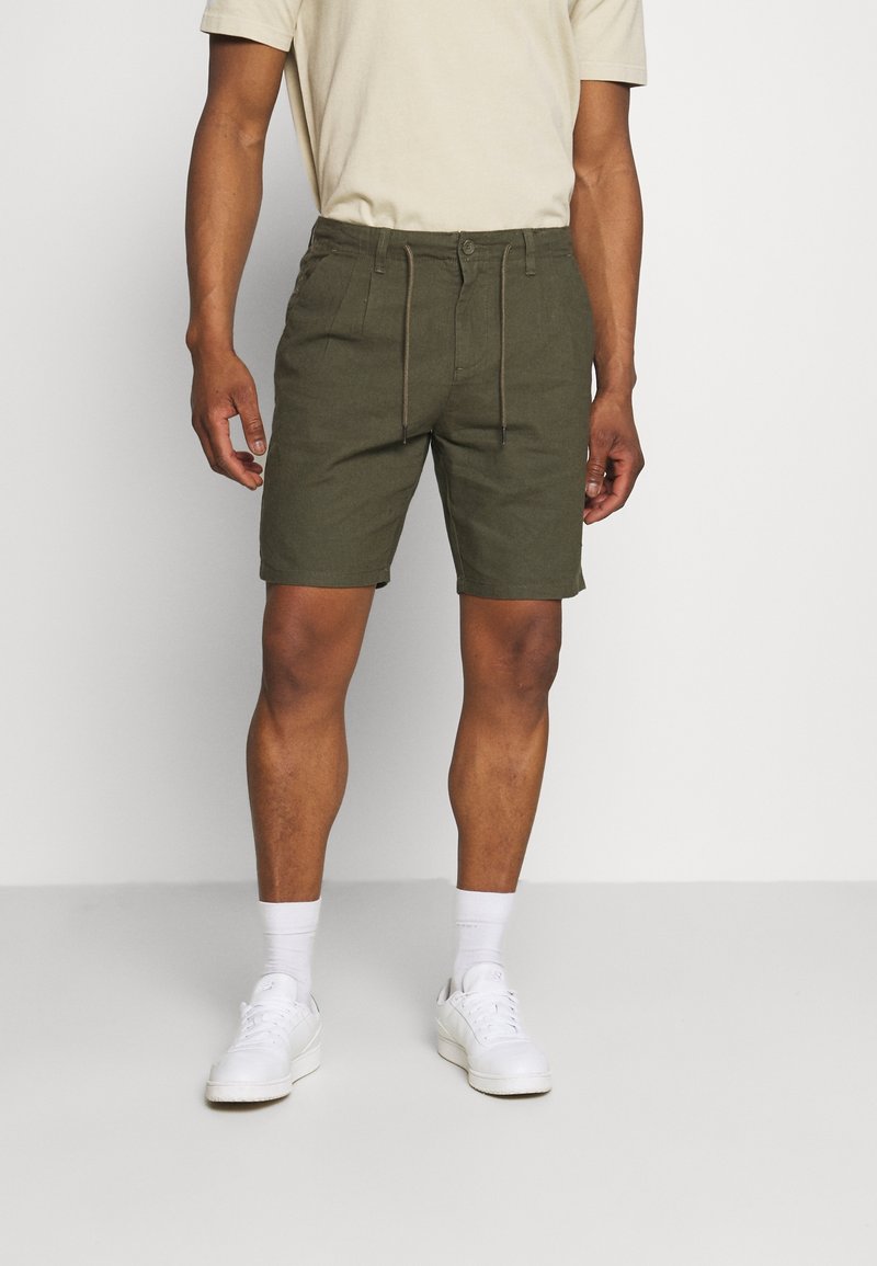 Pantaloni corti verde oliva con cintura regolabile e chiusura a bottone, caratterizzati da una texture di tessuto liscia. Modello che indossa sneakers bianche e calze.