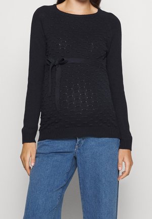 Svart stickad tröja med geometrisk textur, långa ärmar och ett band i midjan. Bärs över blå vida jeans.