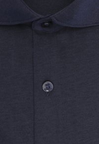 Gros plan d'une chemise en tissu sombre montrant un col et deux boutons noirs, l'un près du col et l'autre en dessous.