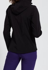 Czarna dopasowana kurtka z kapturem, charakteryzująca się opływowym designem, teksturowanym materiałem i bezszwowym wykończeniem. Połączona z fioletowymi spodniami sportowymi.