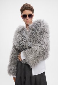 Puszysta, szara kurtka z długimi, teksturowanymi rękawami i kołnierzem, noszona na białej koszuli, w połączeniu z ciemną, plisowaną spódnicą i okularami przeciwsłonecznymi.