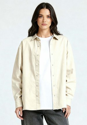 Femme aux longs cheveux bruns portant une chemise beige boutonnée sur un haut blanc et un pantalon gris foncé, debout devant un fond clair uni.