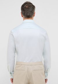 Camicia a maniche lunghe di colore azzurro chiaro con polsini abbottonati, caratterizzata da una texture liscia. Abbinata a pantaloni beige, vista di spalle.