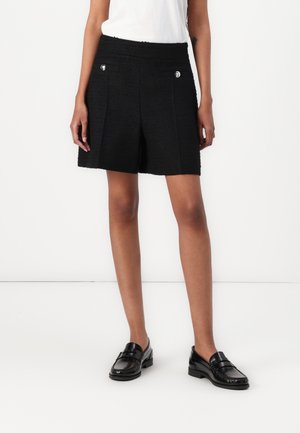 Femme portant un short texturé noir avec des poches avant boutonnées, un haut blanc et des mocassins en cuir noir, debout sur fond blanc.