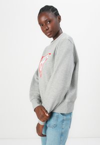 Grijze sweatshirt met een rood en wit logo op de voorkant, ronde halslijn, geribbelde boorden en zoom; gecombineerd met lichtblauwe jeans.