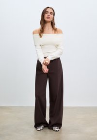 Weißes, geripptes Oberteil mit schulterfreiem Schnitt, kombiniert mit dunkelbraunen, weit geschnittenen Hosen und gemusterten Schuhen. Einfaches Design, weicher Stoff.