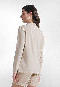 Top beige a maniche lunghe con scollo rotondo, tessuto morbido e dettagli cuciture visibili. Indossato con pantaloncini beige abbinati, con una vestibilità rilassata.