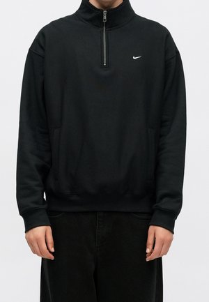 Osoba ubrana w czarną bluzę Nike z suwakiem do ćwiartki długości, długimi rękawami i kieszeniami, stojąca na gładkim jasnym tle.