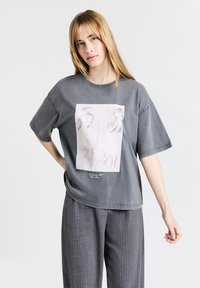 T-shirt oversize gris en coton, avec un imprimé à l'avant représentant deux danseurs dans un style de croquis léger. Texture douce et coupe décontractée.