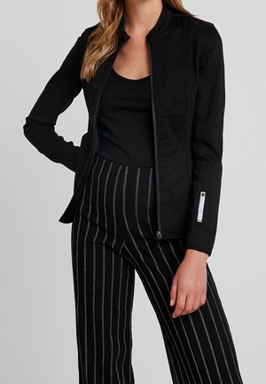 Veste zippée noire avec un col haut, des manches longues et une coupe slim. Présente un léger lustre et une poche zippée sur la manche.