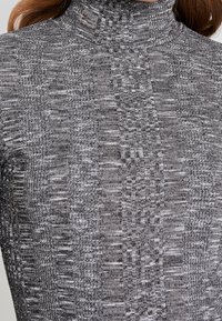 Harmaa neulottu poolopaita, jossa on teksturoitu kuvio ja eri paksuisia lankoja. Ei näkyvää metalliosaa tai koristeita. Pehmeä, vartalonmyötäinen malli.