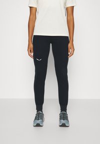 Mørk marineblå joggebukser med logo i hvitt, laget av mykt stoff. Kombinert med grå treningssko med svarte og røde detaljer.