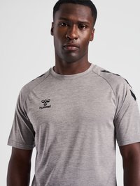 Férfi, szürke hummel sport pólót visel fekete logóval a mellkasán és halszálka mintával az ujjakon, egyszínű háttér előtt áll.