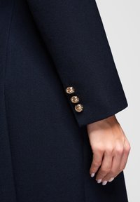 Manche d'un manteau en laine bleu marine avec tissu texturé et quatre boutons décoratifs dorés, mettant en valeur une couleur riche et des détails sur le poignet.