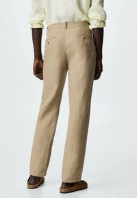 Pantalon en lin beige clair à coupe droite, avec des poches arrière à boutons, et une coupe décontractée. Porté avec des sandales marron.