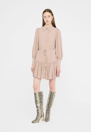 Robe chemise rose clair avec fermeture à boutons, manches bouffantes et taille ajustable par cordon. Associée à des bottes à talons en imprimé serpent, arrivant jusqu'aux genoux.