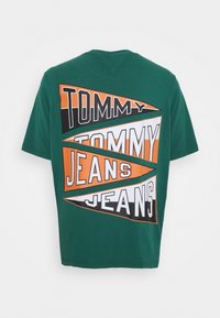 Zelené bavlněné tričko s velkým grafickým designem, na kterém je text "TOMMY JEANS" v tučných, barevných trojúhelnících a pruzích na zádech.
