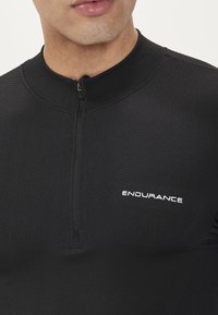 Svart sportströja med hög krage, halvdragkedja, texturerad tygkvalitet och vit "ENDURANCE"-logotyp på bröstet.