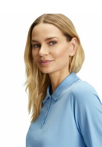 Polo azzurro chiaro con colletto, caratterizzato da un tessuto testurizzato e tre bottoni al collo. Il modello ha capelli biondi lunghezza spalla.